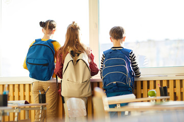 Rear view of dreamy school friends with satchels on backs looking out window while waiting for parents in classroom - obrazy, fototapety, plakaty
