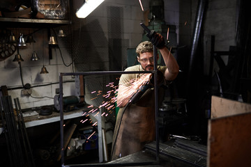 Serious young worker in protective goggles making metal frame and polishing it with sander at table