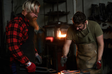 Serious brutal manual workers in aprons hammering hot iron while changing shape of heated metal together in workshop