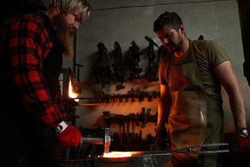 Serious concentrated coworkers in workwear changing shape of heated metal on anvil while producing fence in smithery