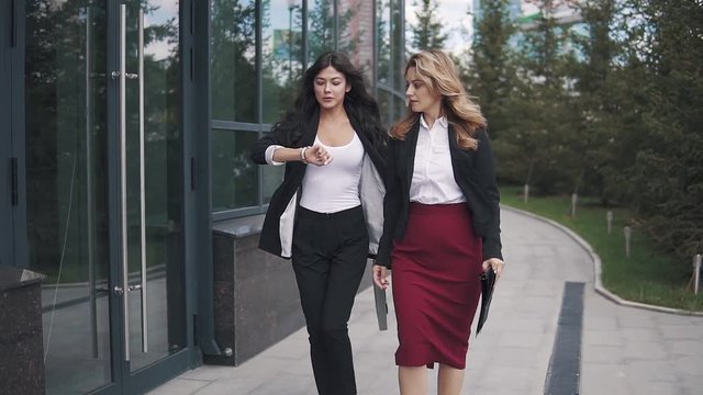 business people go near the office center. two young women in business suits. woman looks at the clock