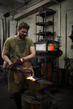 Frowning young male blacksmith in apron and gloves parting metal with cutting torch while standing at anvil in workshop