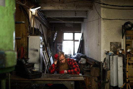 Unhappy exhausted male smith in checkered shirt sitting at table and leaning on hand while trying to sleep in tipped-over workshop