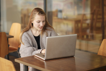 Pretty female student with cute smile keyboarding something on net-book while relaxing after lectures in University, beautiful happy woman working on laptop computer during coffee break in cafe bar