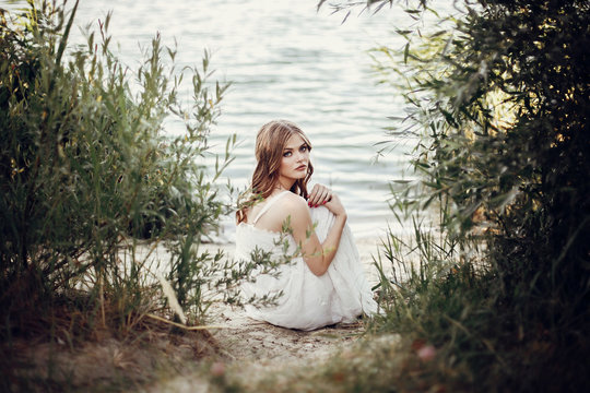 beautiful boho girl sitting on sandy beach at sunset light near lake. attractive young woman in white bohemian dress with windy hair relaxing  near water among green leaves. summer vacation