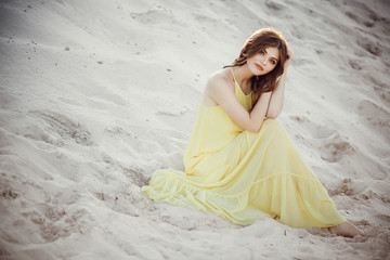 Fototapeta na wymiar beautiful boho girl posing on sandy beach at sunset light near lake. attractive young woman sitting in yellow dress with windy hair and relaxing. calm moment. summer vacation