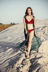 beautiful boho girl in red swimsuit walking on sandy beach at sunset light. attractive young sexy woman in bohemian look with windy hair, amazing eyes and lips. summer vacation