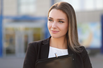 Young beautiful girl in the street next to the building in a jacket and with a notebook.