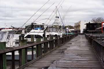 A wooden boardwalk in a small town with a row of sport fishing boats on the river.