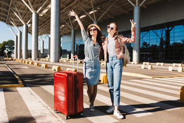 Two cheerful female friends in sunglasses happily looking in camera raising hands up with red suitcase and backpack on shoulder near airport - obrazy, fototapety, plakaty