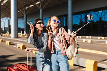 Two cheerful girls in sunglasses while happily showing two fingers gesture taking photo on cellphone together with suitcase and backpack on shoulder outdoor near airport