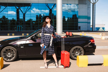 Pretty smiling girl in sunglasses and black hat holding on heels passport with flight ticket and red suitcase near airport with cabriolet on background