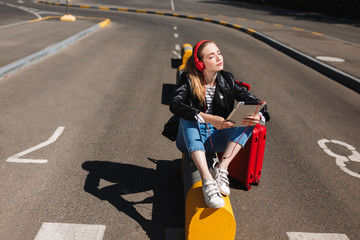 Cute girl in headphones listening music dreamily closing eyes with tablet in hand and suitcase near on road of airport