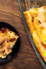 Cottage cheese casserole in a large glass baking dish and one serving on a round plate, top view on a wooden background.