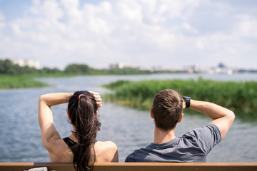 High section back view of young couple sitting on bench enjoying lake view in park, copy space