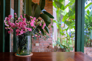 Pink carnation in a clear glass vase on a wooden table in a cafe with green plants in background