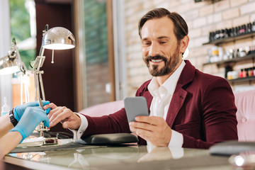 Meanwhile nail bar. Low angle of joyful mature man getting nails done and using phone