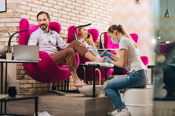 Pedicure for men. Jolly mature man getting toenails done and using laptop