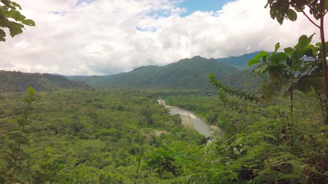 A high-angle view of the meandering Mulatos River cutting through a dense green forest valley near Tena in the Ecuadorian Amazon, foliage framing it and mountains under a cloudy sky.