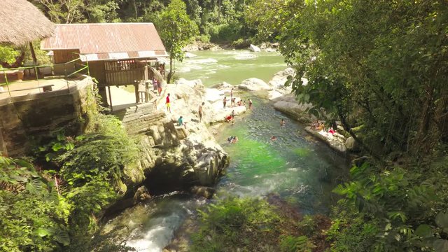 A high-angle view of the Mulatos River with a natural pool where people swim among large rocks at Tena Laguna Azul in the Ecuadorian Amazon, a wooden riverside structure with a corrugated roof.
