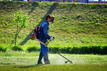 Unknown gardener in uniform mowing the grass with lawn mower in the park. Worker wears Personal...