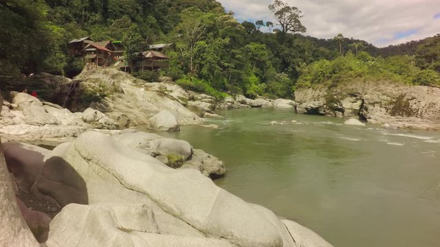 A wide shot panning across the fast-flowing Mulatos River in a lush tropical valley near Tena in the Ecuadorian Amazon, large smooth boulders and traditional wooden huts on the rocky bank.