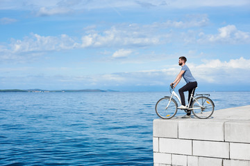 Fototapeta premium Young bearded man tourist on bicycle on high paved stone sidewalk enjoying clear blue sea water. Active holiday and tourism concept.