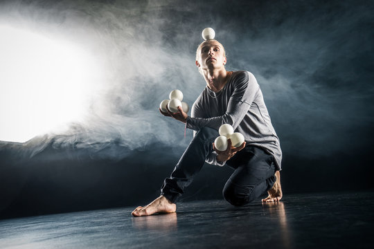 Blond juggler sitting on the floor with white balls on black background