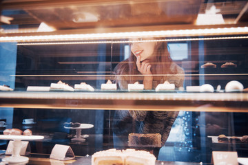 Smiling woman at camera through the showcase with sweet and cakes in modern cafe interior