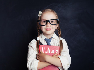 Happy little child girl hugging italian book against chalkboard background in school classroom....