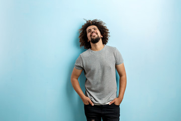 A curly-headed handsome man wearing a gray T-shirt and ripped jeans is standing and smiling with his hands in the pockets, looking upwards over the blue background.