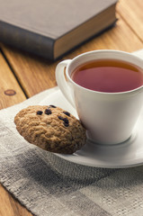 A cup of tea and some chocolate chip cookies over a books on a brown wooden table.