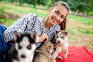 A beautiful smiling woman with a ponytail and wearing a striped shirt is cuddling with  three sweet husky puppies while resting on the red blanket on the lawn. Love and care for pets.
