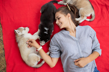 A beautiful smiling woman with a ponytail and wearing a striped shirt is cuddling with  three sweet husky puppies while resting on the red blanket on the lawn. Love and care for pets.