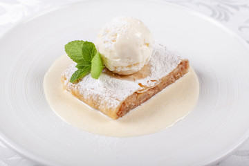 Fruit apple strudel cake served with ice cream, mint leaf and vanilla sauce. Classical austrian dessert on white plate. Sweet dessert in the restaurant. Morning light.