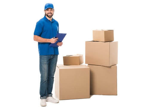full length view of handsome young delivery worker holding clipboard and smiling at camera while standing near boxes isolated on white