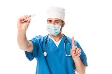 doctor in medical mask holding thermometer, pointing up with finger and looking at camera isolated on white