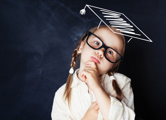 Pensive kid schoolgirl wearing graduation hat in school classroom. School concept.