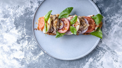 Delicious Italian sandwich with smoked chicken, mozzarella, tomatoes, sweet onions and basil leaves on a round plate on a concrete background. Top view