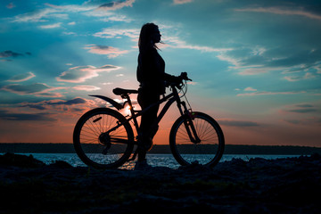 Naklejka na ściany i meble Silhouette of a young woman with a bicycle on the lake at sunset