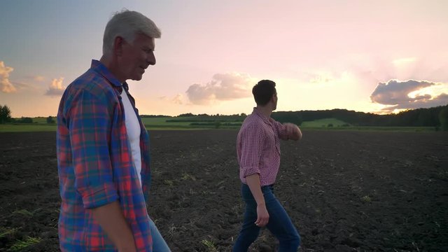 Side view of old father and adult son walking on cultivated field, beautiful landscape with sunset in background