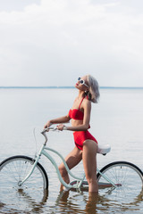 Fototapeta premium beautiful young woman in red trendy bikini sitting on bicycle in water