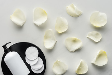 top view of bottle of cream and cosmetic pads on plate, scattered rose petals on tabletop, beauty concept