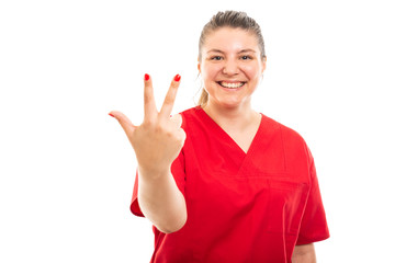 Young medical nurse wearing red scrub showing number three gesture.