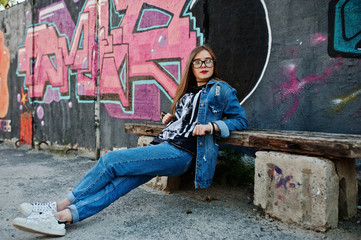 Stylish casual hipster girl in jeans wear and glasses against large graffiti wall.