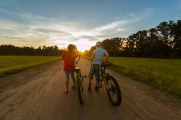 Obraz premium childs riding one bike together on sunny summer evening. sitting on bicycle rack. Family of two people enjoying traveling in scenic field over sunset sky background.