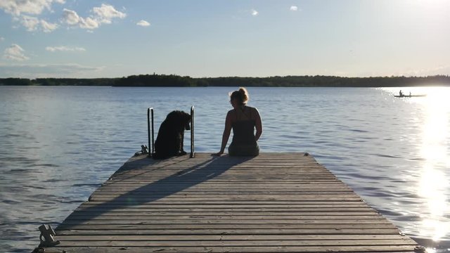 Woman sitting on the end of the dock with dog staring at the water. She tries to get the dog to sit closer to her.