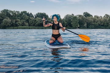attractive tattooed girl with blue hair surfing on paddle board in water