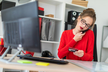 A young girl is sitting at the desk in the office, holding a bank card in her hand, typing on the...