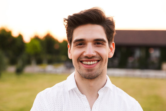 Photo closeup of cheerful young man wearing white shirt smiling, while walking outdoor through green field in countryside - Powered by Adobe
